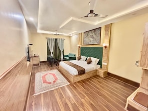 A hotel room with a wooden floor, a bed with a green headboard, a ceiling fan, and a wall-mounted artwork. - KRIMZON MANOR (Srinagar)