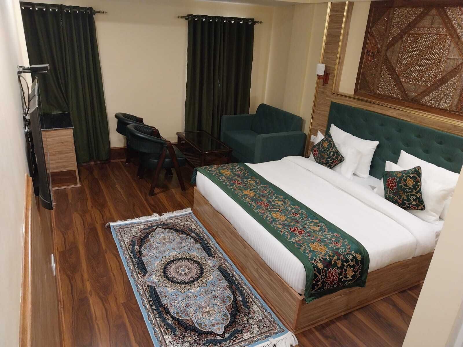 Quarto de hotel com cama, duas cadeiras, escrivaninha, televisão e um objeto decorativo na parede.