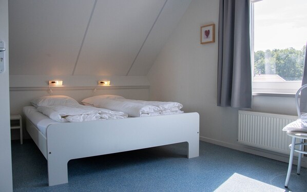 Ett sovrum med en vit säng, två sänglampor, ett element och ett fönster med gardiner.