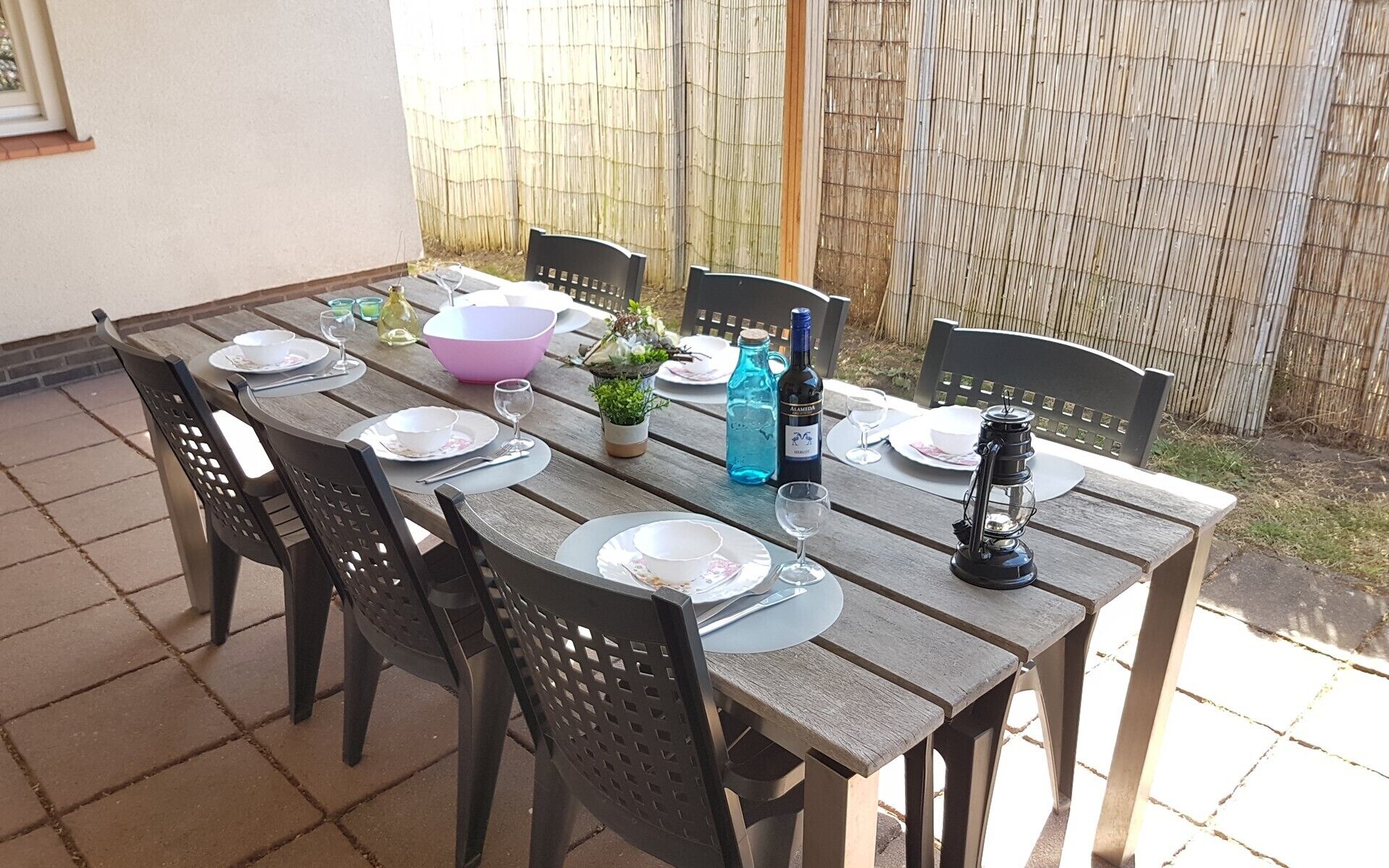Mesa de madeira posta para seis lugares, com cadeiras, uma garrafa de vinho, um candeeiro e um vaso de plantas.