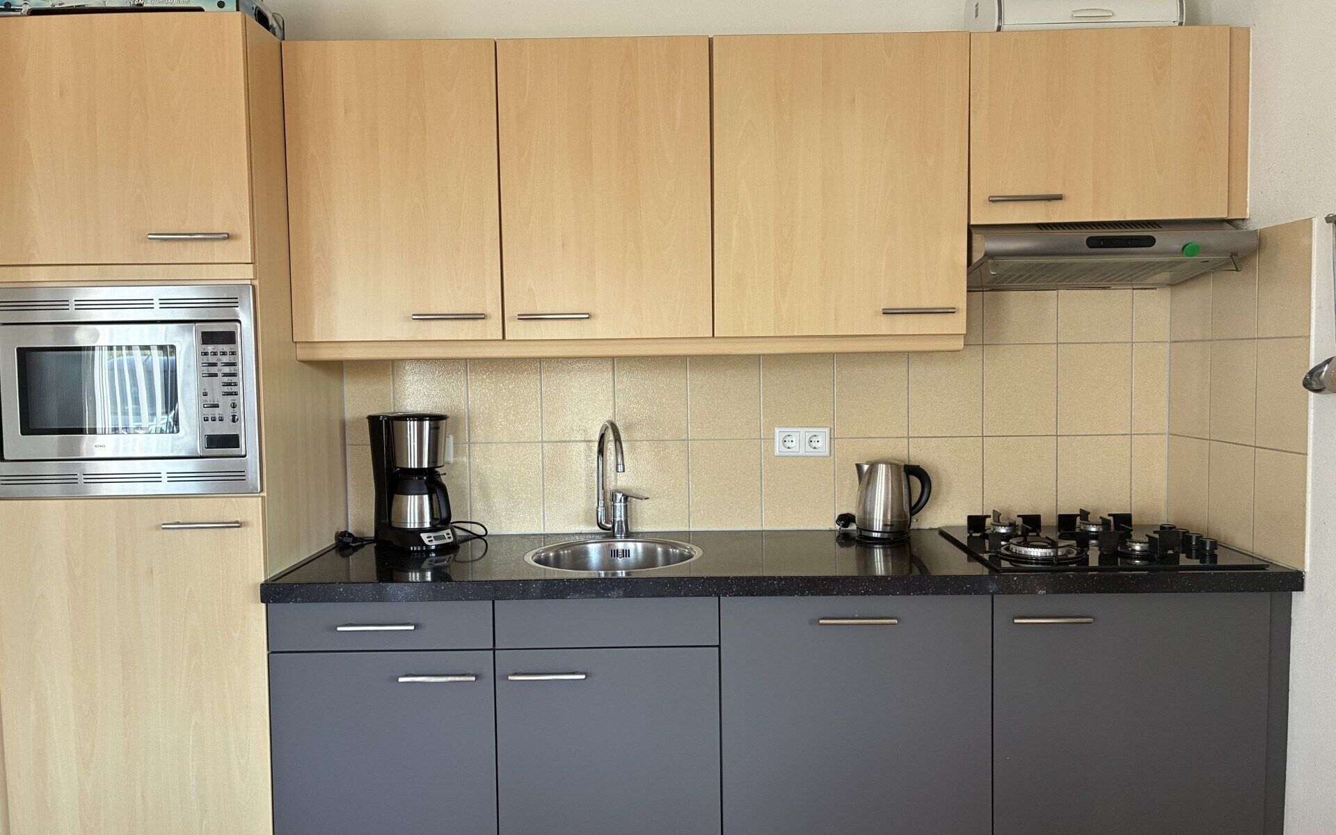 Een keuken met houten kastjes, een roestvrijstalen magnetron, een koffiezetapparaat, een waterkoker en een gasfornuis.