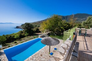 A swimming pool with a stone deck, a thatched umbrella, and a view of the sea and hills. - Villa Pelagos Kyma in Lefkada (Lefkada)