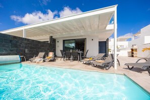 A modern outdoor pool area with lounge chairs, a covered seating area, and a stone wall. - Villa Gara 8 in Estepona (Yaiza)