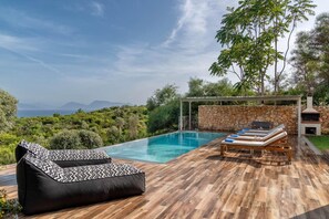 A poolside area with a wooden deck, a black and white patterned lounge chair, and a stone wall. - Villa Melanthi in Lefkada (Lefkada)