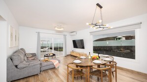 A modern living room with a sofa, dining table, and chairs, a TV, and a large window overlooking a body of water. - Villa Yaiza Secret 1 (Yaiza)