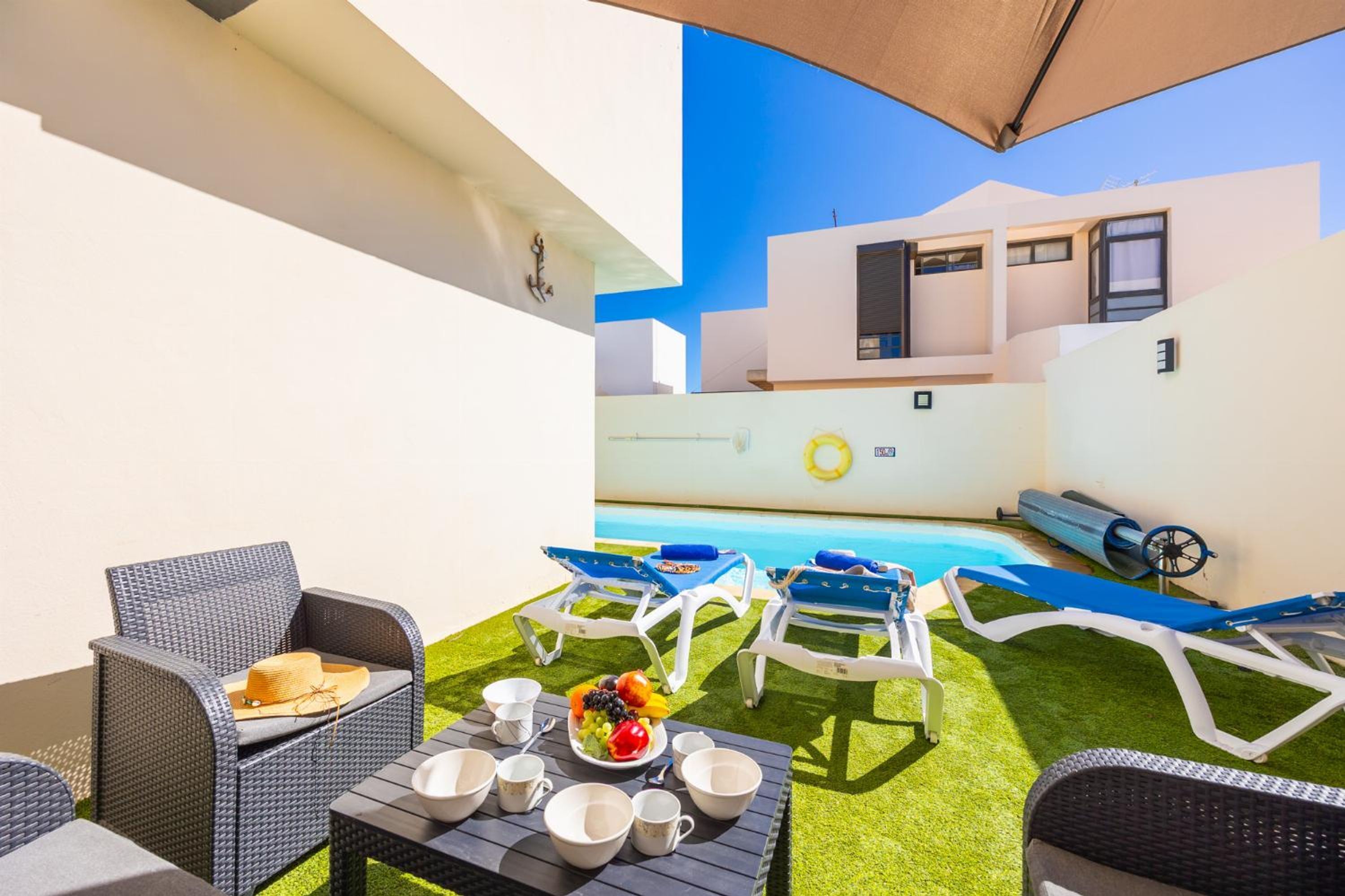 Zona de piscina amb gandules, una taula amb un plat de fruita i un edifici blanc al fons.