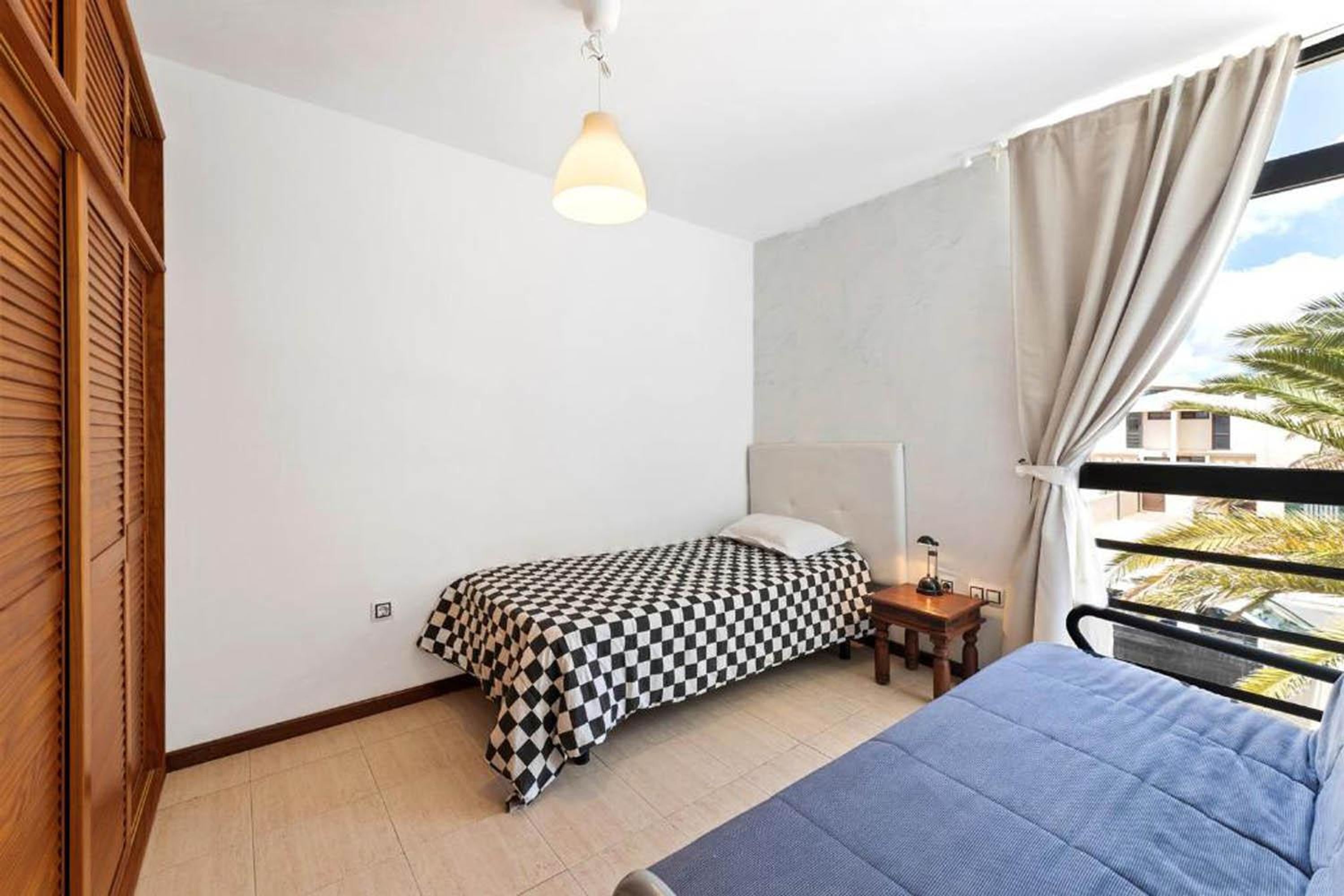 A bedroom with a checkered bedspread, a bedside table with a lamp, a window with curtains, and a view of palm trees.