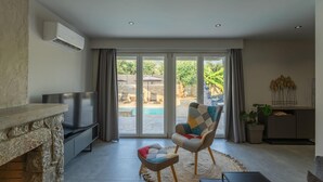 A living room with a stone fireplace, a flat-screen TV, a comfortable armchair, a small ottoman, and a view of a swimming pool through large sliding glass doors. - Villa Omega in Alonnisos (West Mani)