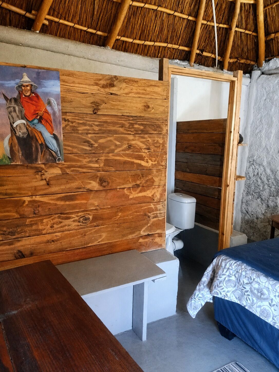 A room with a wooden wall, a bed with patterned bedding, a bench, a toilet, and a painting of a person on a horse.