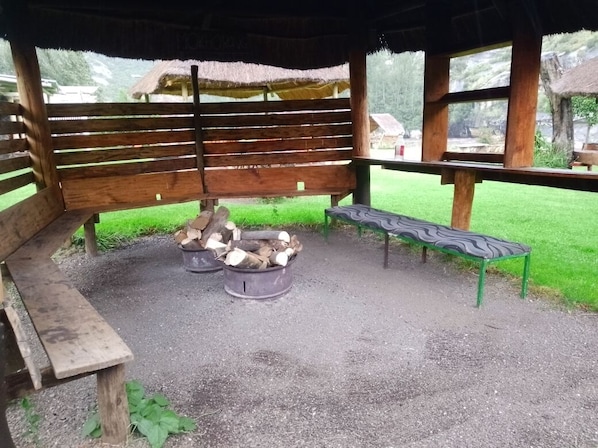 A wooden bench, a metal fire pit with logs, and a green bench under a shelter.