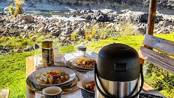 Een tafel buiten gedekt met eten, een thermoskan en een kopje, met uitzicht op een rotsachtig landschap en een wateroppervlak.