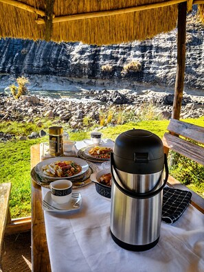 Mesa al aire libre con comida, una termo y una taza, con vista a un paisaje rocoso y un cuerpo de agua.