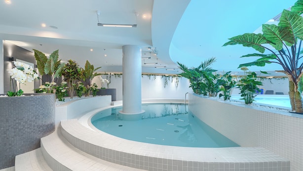 A modern indoor pool area with a curved design, surrounded by tiled walls and steps, and decorated with various plants and flowers. - MARGI Milky Apartament (Szczecin)