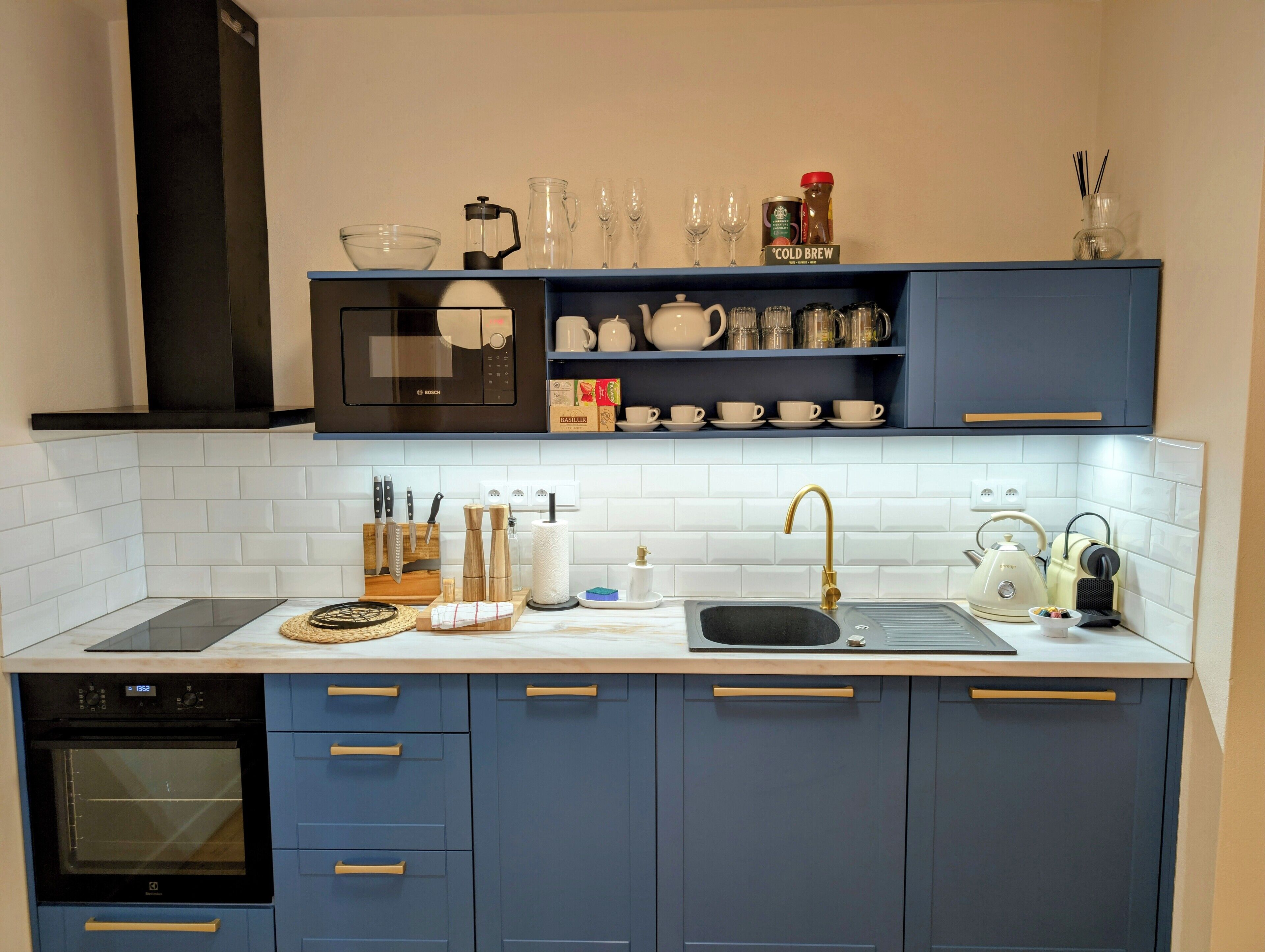 A modern kitchen with blue cabinets, a Bosch microwave, a sink with a gold faucet, and a coffee maker.