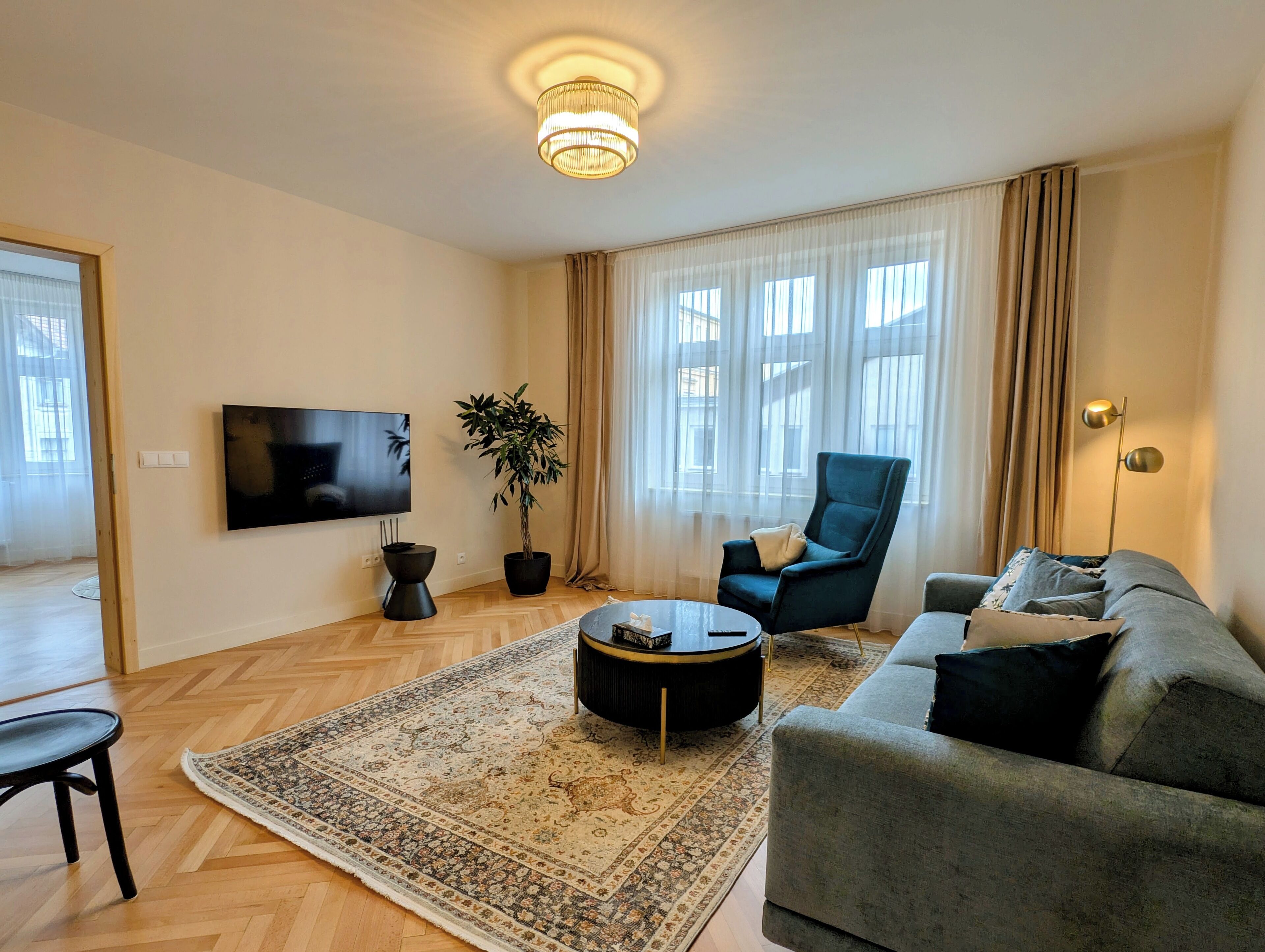 Ein modernes Wohnzimmer mit Flachbildfernseher, einer Topfpflanze, einem blauen Sessel, einem grauen Sofa und einem gemusterten Teppich.