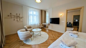 Hotellihuoneessa on sänky, kaksi nojatuolia, pieni pyöreä pöytä, televisio ja työpöytä tuolilla.