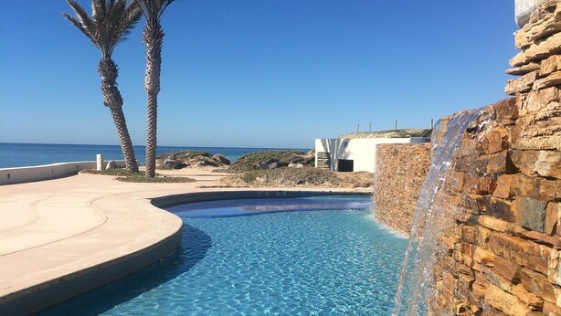 A clear blue pool with a stone wall on one side and a palm tree on the other. - Tessoro 704 - Amazing beachy ocean front condo (Puerto Peñasco)