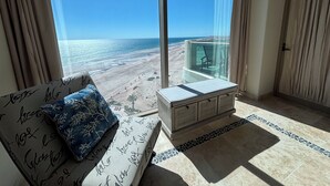 A room with a large window overlooking a beach, a sofa with patterned cushions, a glass-top table, and a tiled floor. - Tessoro 704 - Amazing beachy ocean front condo (Puerto Peñasco)