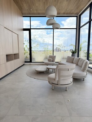 Un salon moderne avec un canapé d’angle, une table basse et un téléviseur fixé à un mur en bois. De grandes fenêtres offrent une vue sur le paysage urbain.