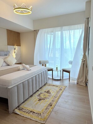 Une chambre à coucher moderne, dotée d’un grand lit, d’une table de chevet, d’une chaise et offrant une vue sur l’extérieur à travers des rideaux légers.