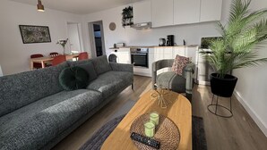 A modern living room with a grey sofa, a wooden coffee table with candles, a remote control, and a kitchen in the background. - Stranddistel 5 - "Stranddistel" vacation home with 5 residential units... (Dahme)