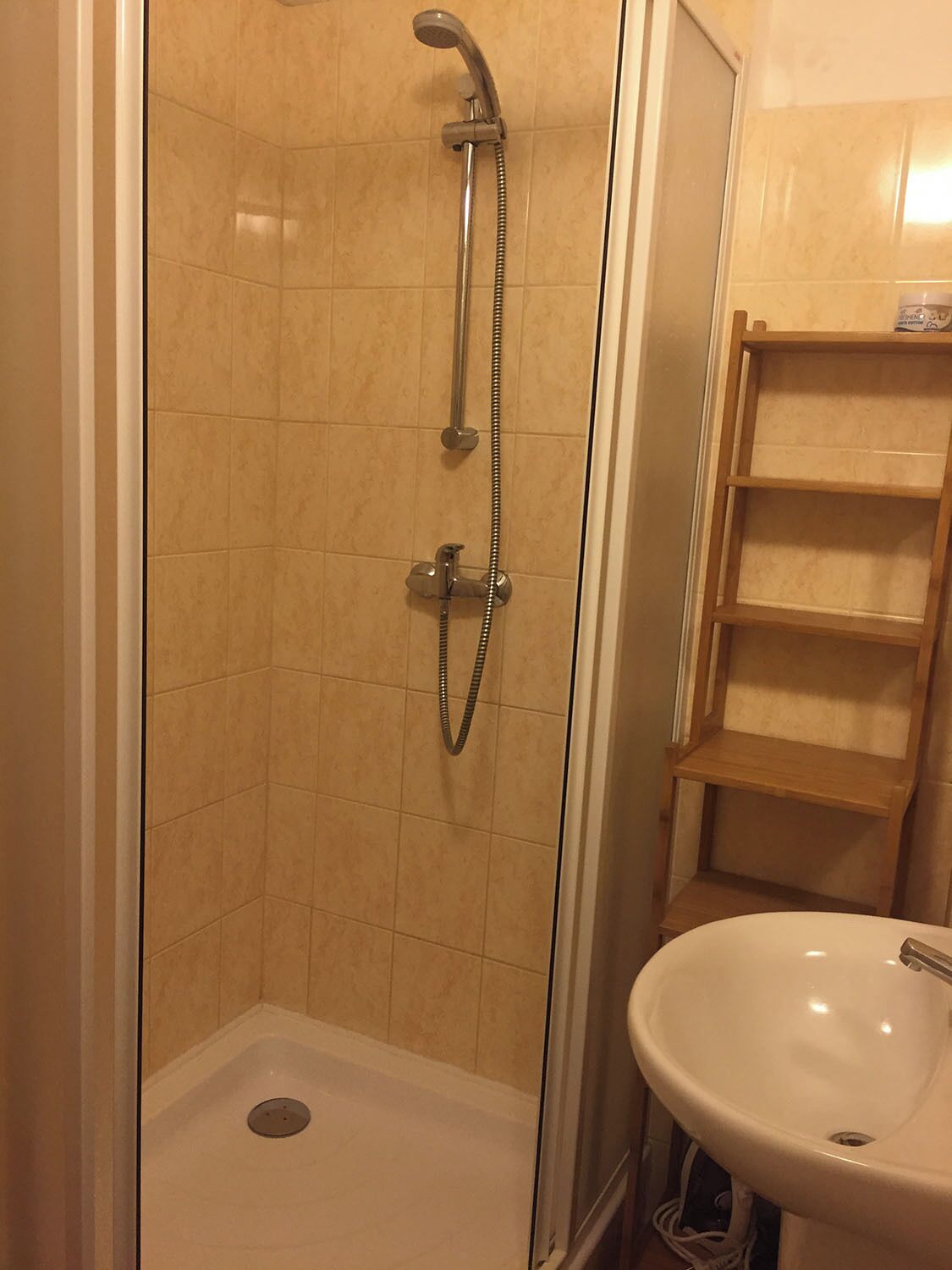 Eine Dusche mit Handbrause und Wandarmatur, ein weißes Waschbecken mit Einzelbecken sowie eine Holzregaleinrichtung in einem Badezimmer mit beigen Fliesen.