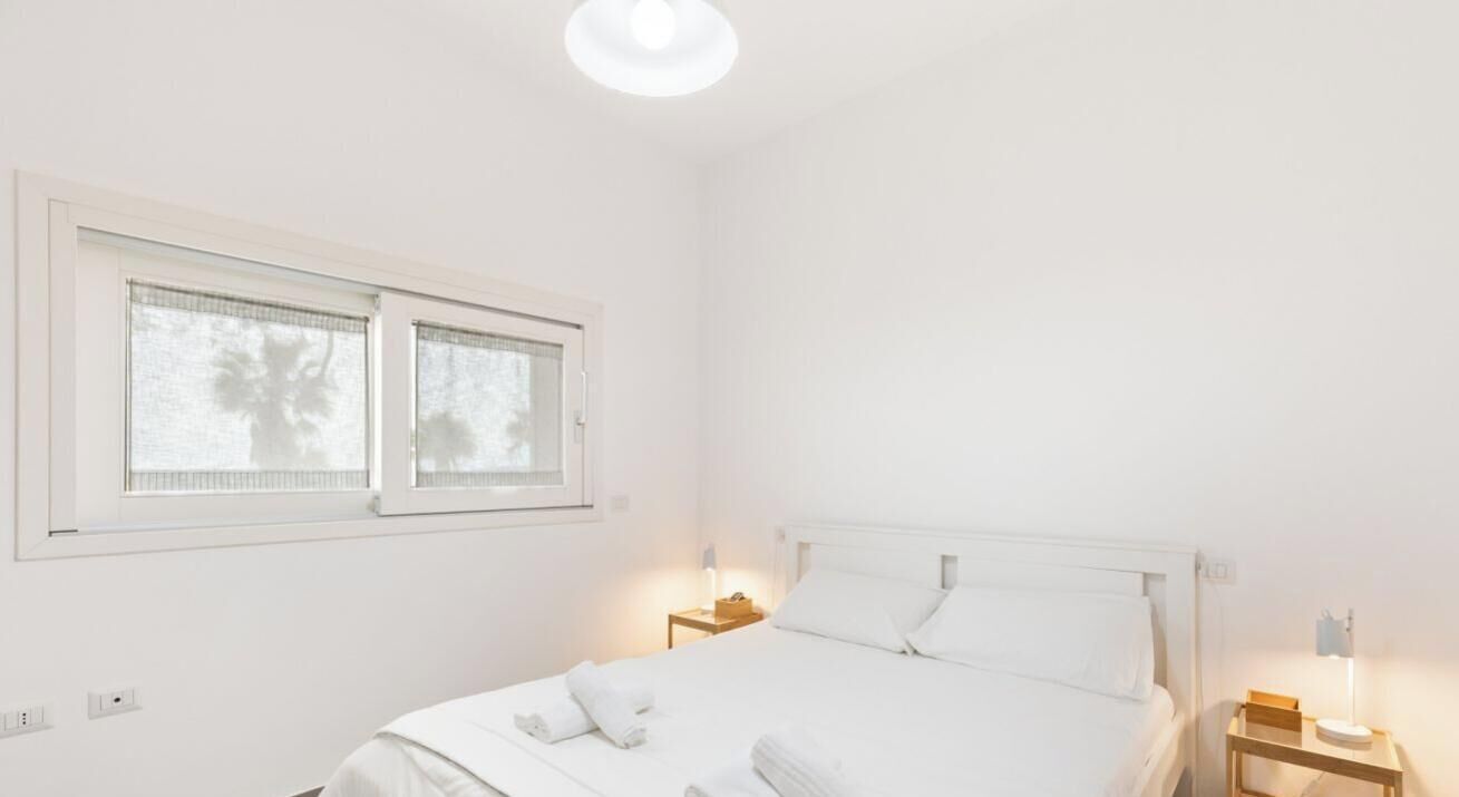 A white room with a bed, two bedside tables, a window, and a ceiling light.