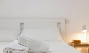 Una cama bien tendida con sábanas blancas, una lámpara de noche y una mesita de madera.