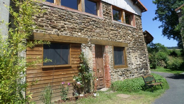 Un bâtiment en pierre et en bois, avec une porte d’entrée en bois, de grandes fenêtres et un banc à l’extérieur.