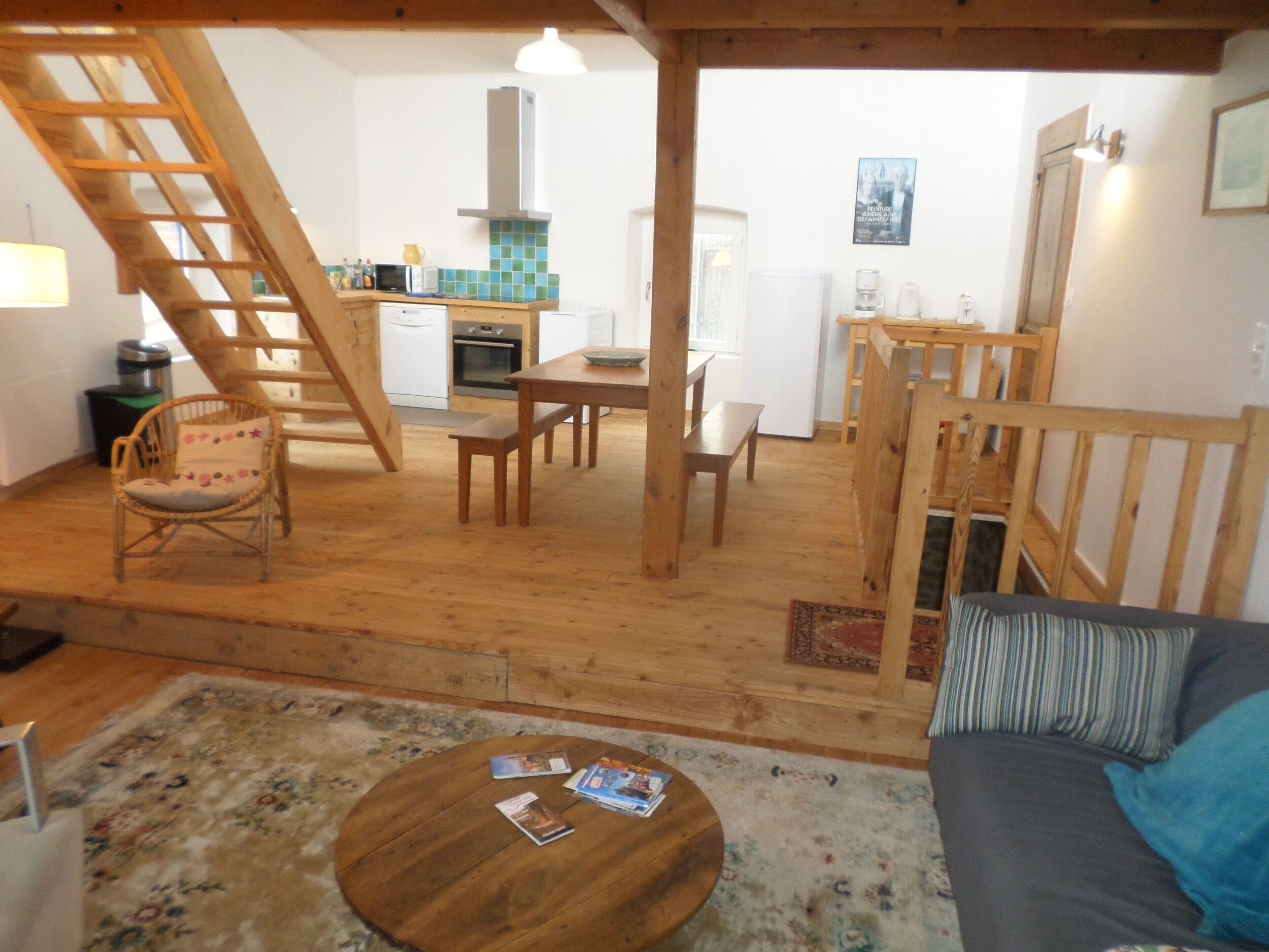 A cozy living area with a wooden floor, a sofa, a coffee table with magazines, a dining table with chairs, a kitchen with a stove and microwave, and a staircase.