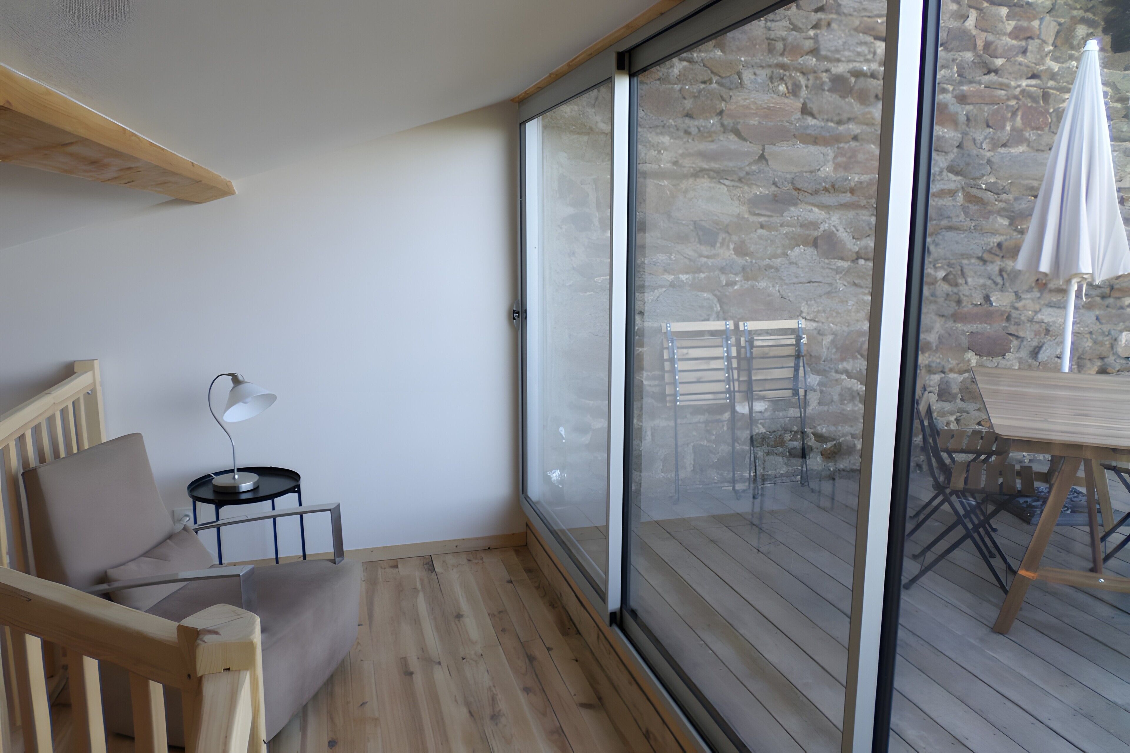 Ein Zimmer mit Holzboden, einer Couch, einem kleinen Tisch mit Lampe und einer Schiebetür aus Glas, die zu einem Außenbereich mit Tisch und Stühlen führt.