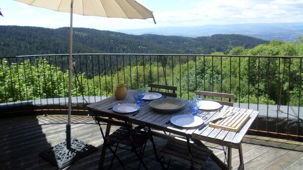 Une terrasse en bois avec une table dressée pour deux personnes, un parasol beige et une vue pittoresque sur une vallée boisée.