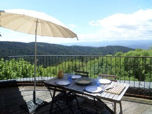 Une terrasse en bois avec une table dressée pour deux personnes, un parasol beige et une vue pittoresque sur une vallée boisée.