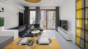Ein modernes Wohnzimmer mit einem schwarzen Ledersofa, einem Esstisch mit Blumenvase, einem Fernseher auf einem Lowboard und einem Balkon mit Blick auf Gebäude.