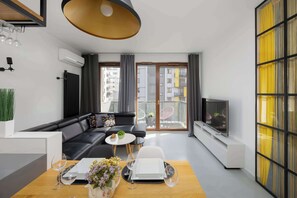 Ett modernt vardagsrum med en svart lädersoffa, ett matbord med en vas med blommor, en tv på ett tv-bord och en balkong med utsikt över byggnader.