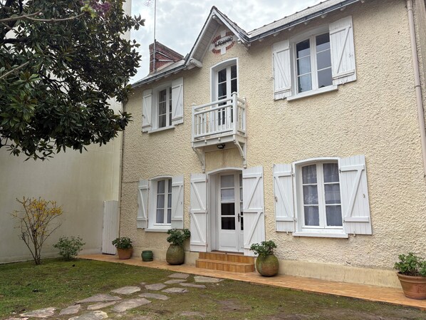 Un immeuble de deux étages avec des volets blancs, un balcon et un petit jardin agrémenté de plantes en pot.