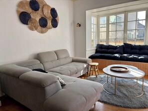 Un salon avec un canapé gris, une table basse ronde et une grande fenêtre donnant sur des immeubles.