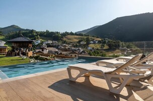 Zona de piscina con sillones de descanso, una terraza de madera y vistas a un pueblo en la ladera.