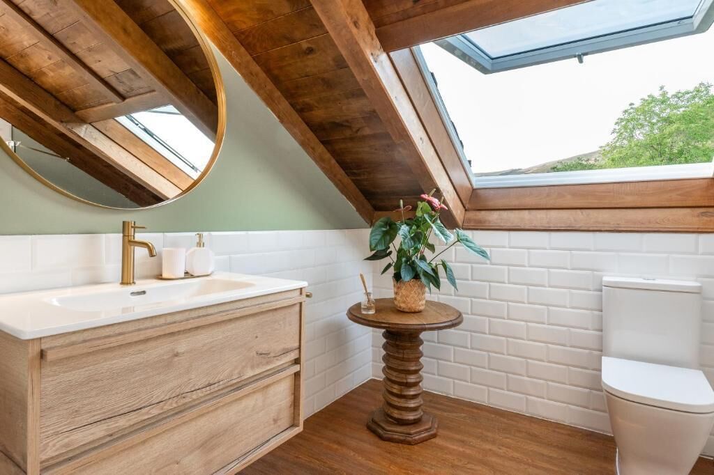 Een badkamer met een houten wastafelmeubel, een rond tafeltje met een plant, een wit toilet en een dakraam.