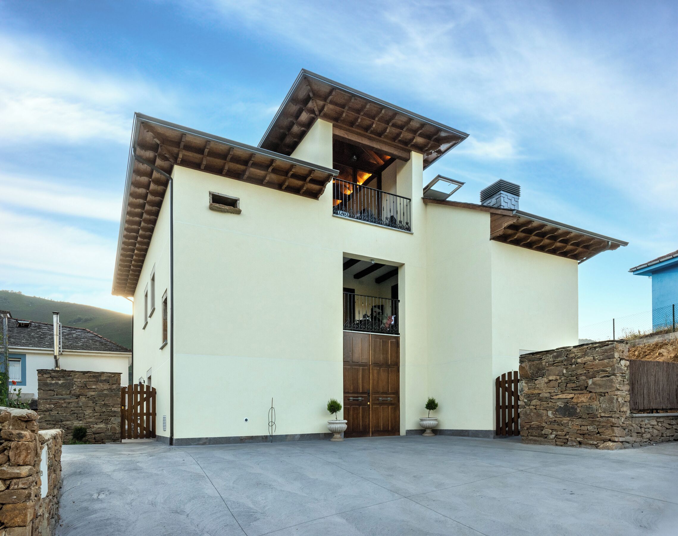 A modern two-story house with a balcony, a stone wall, and a large wooden door.
