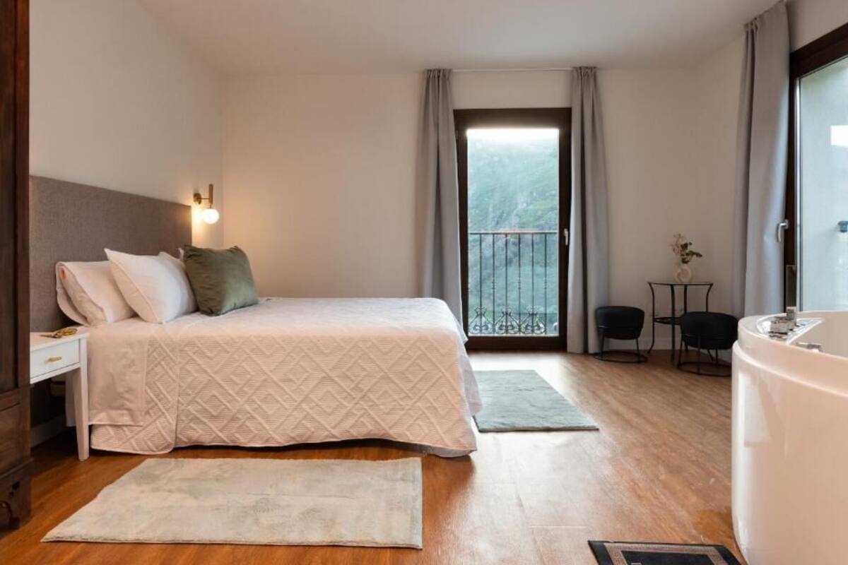 A bedroom with a bed, bedside tables, a small table with chairs, a bathtub, and a balcony with a railing.