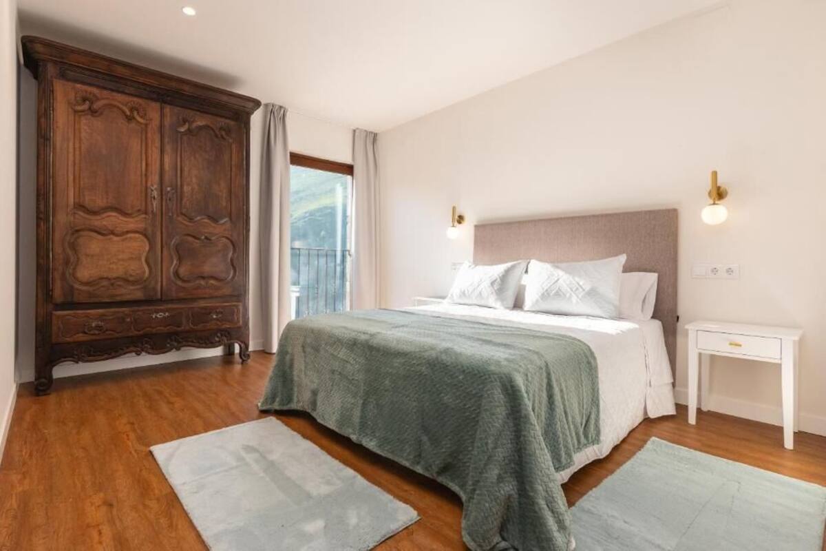 A bedroom with a large wooden wardrobe, a bed with white bedding, a nightstand, and a window with curtains.