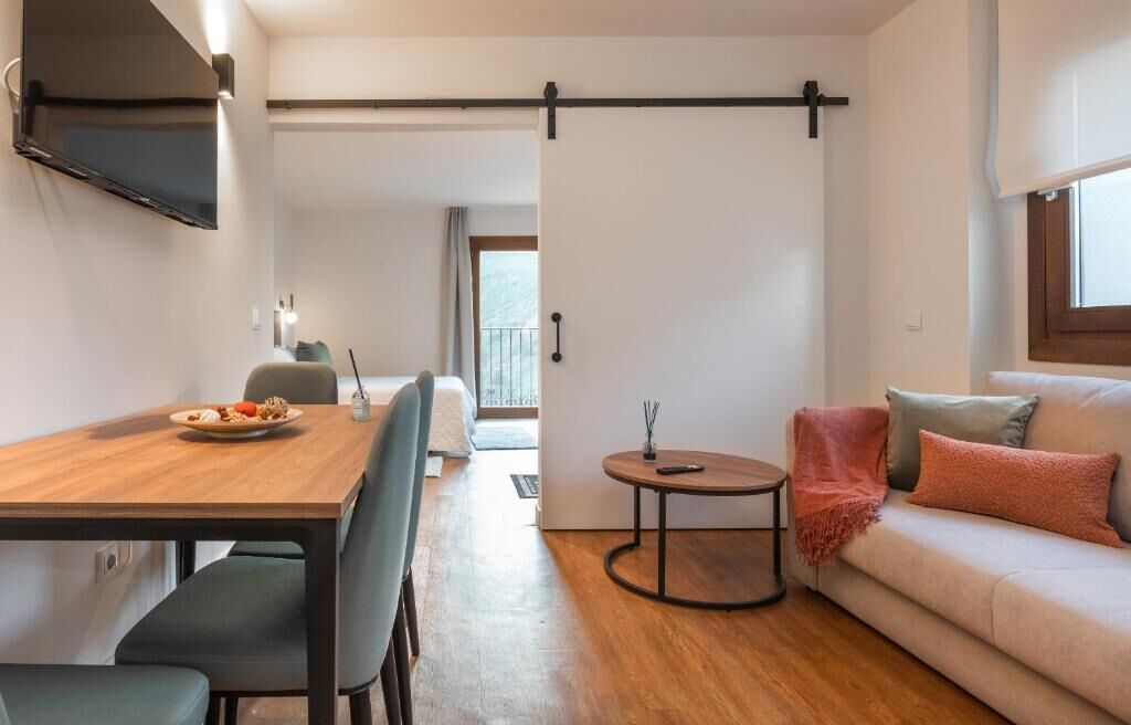 A modern living room with a sofa, a dining table with chairs, a TV mounted on the wall, and a sliding door leading to another room.