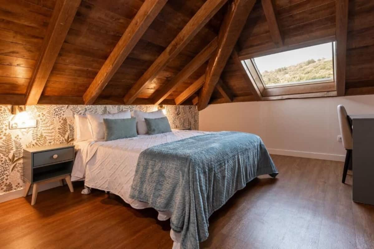 A bedroom with a sloped ceiling, a bed with a blue blanket, a nightstand, and a desk.