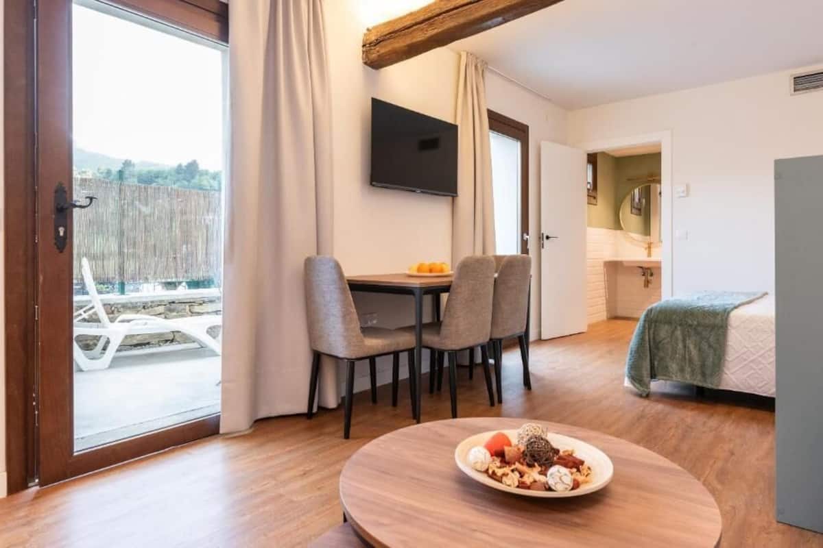 A room with a dining table, four chairs, a television on the wall, a bed with a green blanket, and a view of a bridge and trees outside.