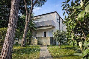 Une maison moderne à deux étages, avec un chemin en béton menant à l’entrée, entourée d’arbres et de verdure.
