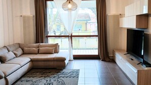 Un salon avec un canapé beige, un téléviseur à écran plat, un tapis à motifs et une vue dégagée sur l’extérieur grâce à une grande fenêtre.