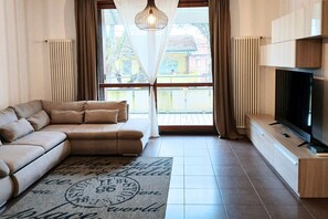 Un soggiorno con un divano color beige, una televisione a schermo piatto, un tappeto decorato e vista attraverso un'ampia finestra.