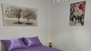 A bedroom with a purple bedspread, a painting of trees, a floral artwork, and a hanging light fixture.