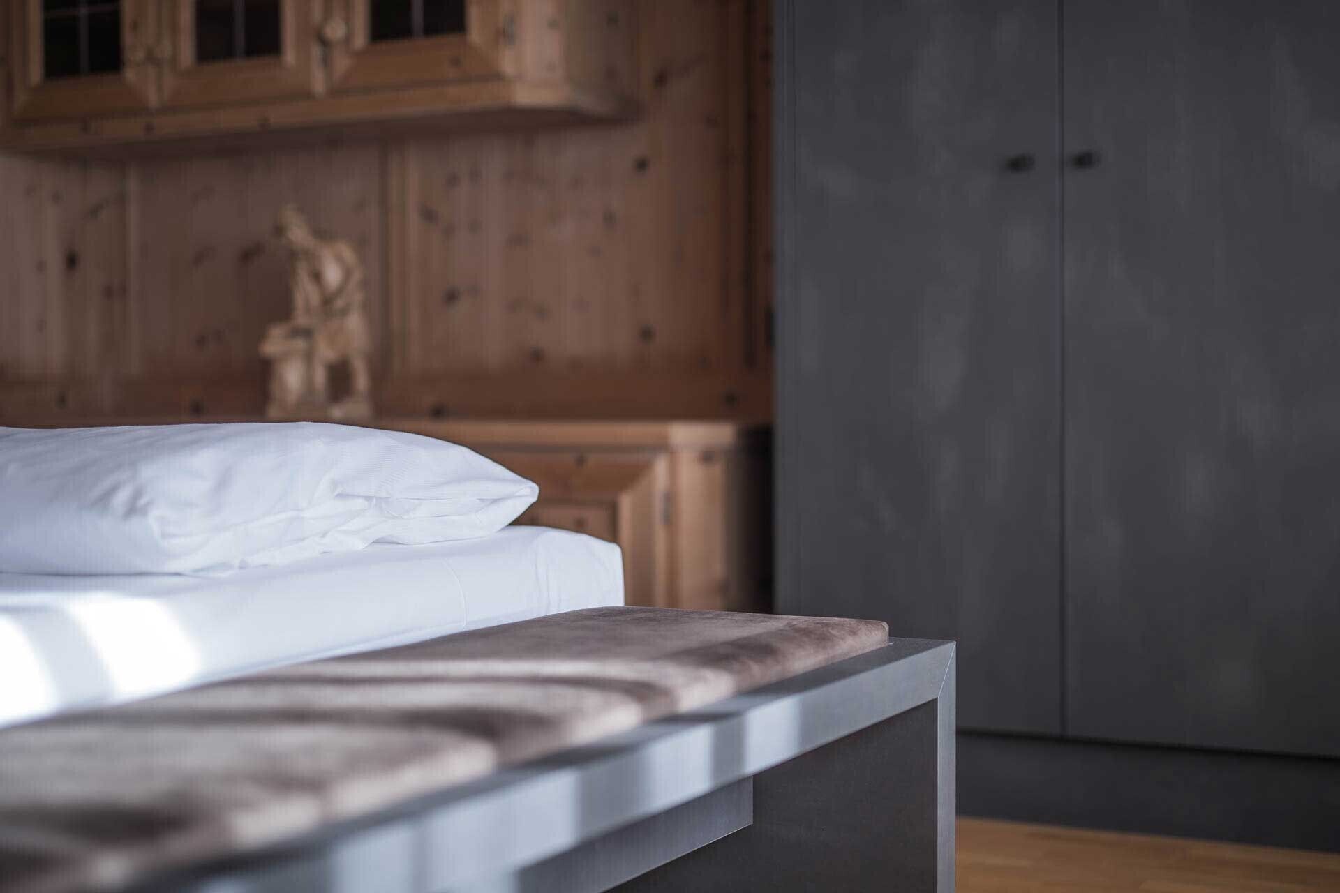 Un lit avec du linge de lit blanc et une tête de lit en bois, dans une pièce aux murs lambrissés et agrémentée d’une commode gris foncé.